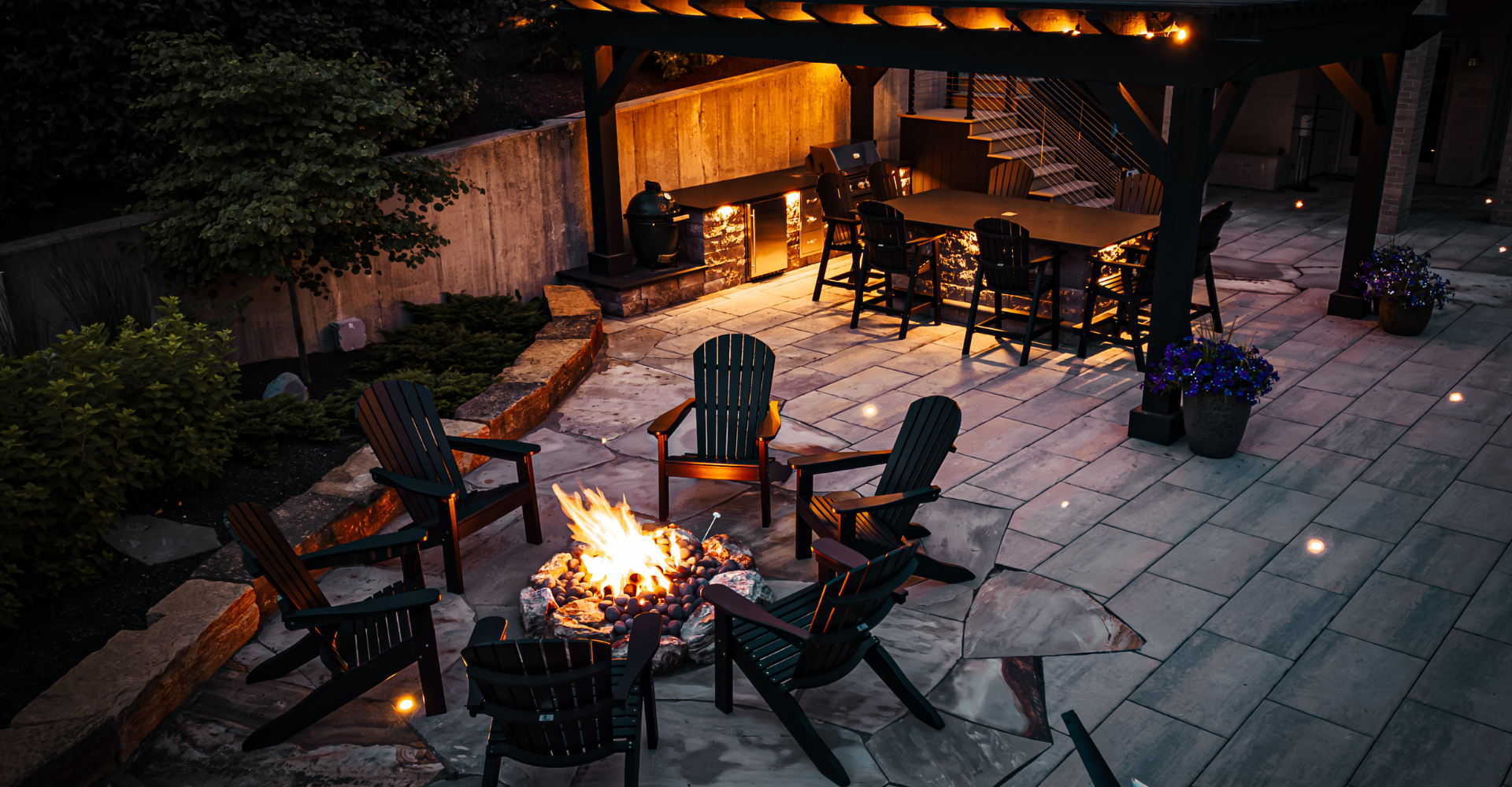 Nighttime closeup view of a firepit and chairs in front of a pergola with outdoor kitchen and dining, as well as lighting