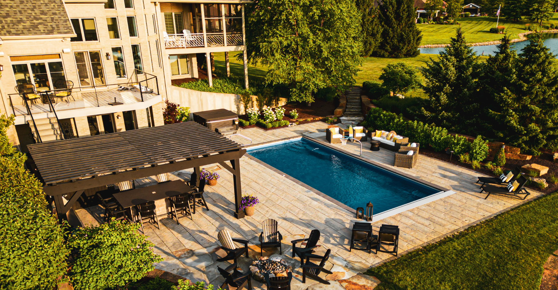 Aerial sunset view of a design build featuring a pergola with outdoor kitchen and dining, firepit, pool, and furniture surrounded by softscaping