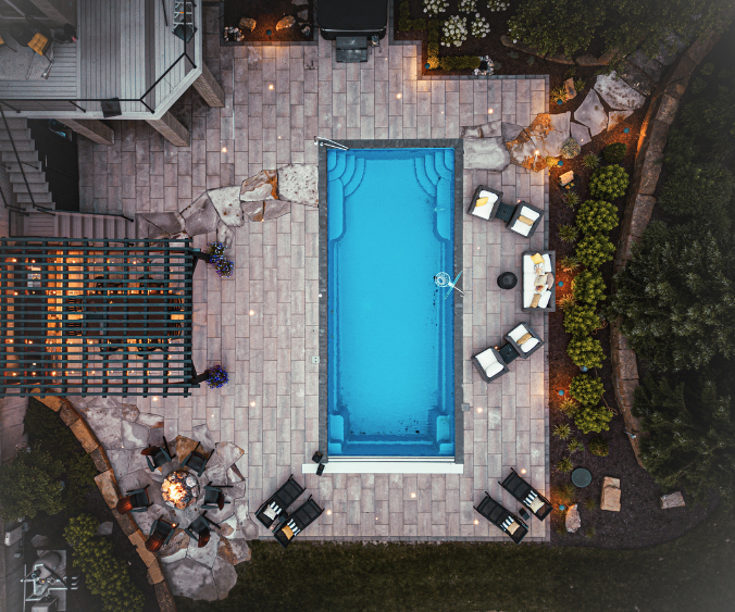 An aerial dusk view of a design build featuring a patio with embedded lighting, pool, firepit, and pergola with outdoor kitchen and dining