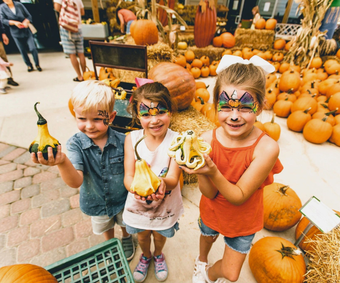 Three children with faces panted, holding uniquely shaped gourds amongst fall decorations.